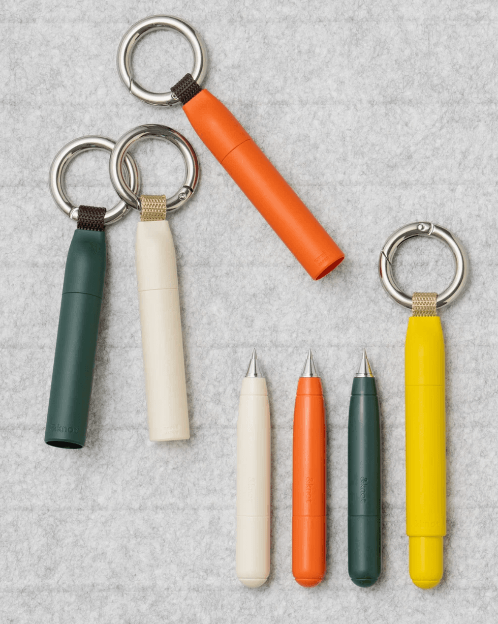 Colorful keychain pens in green, cream, orange, and yellow on a gray background.