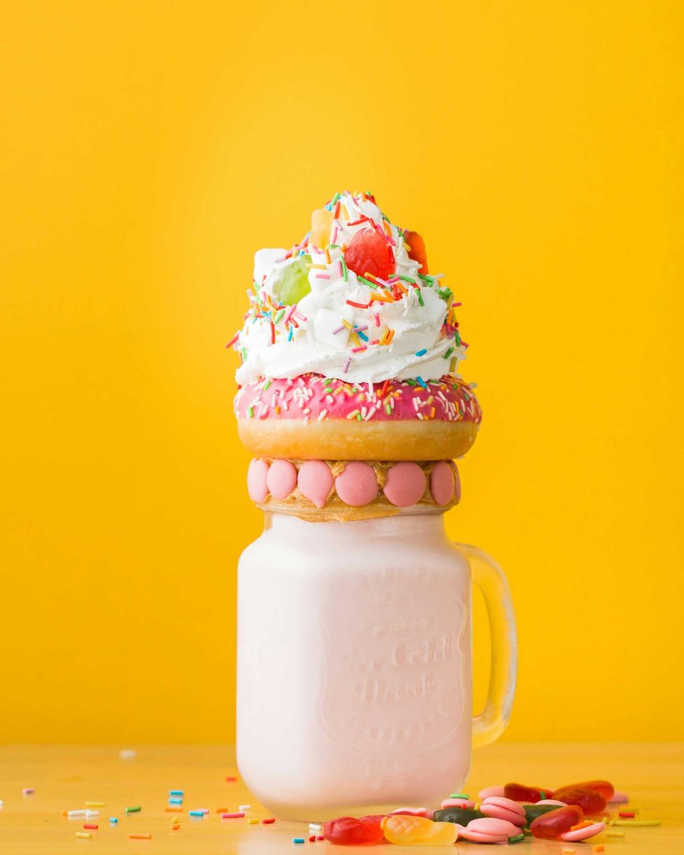 Colorful milkshake with donut, whipped cream, and sprinkles in a mason jar on yellow background.