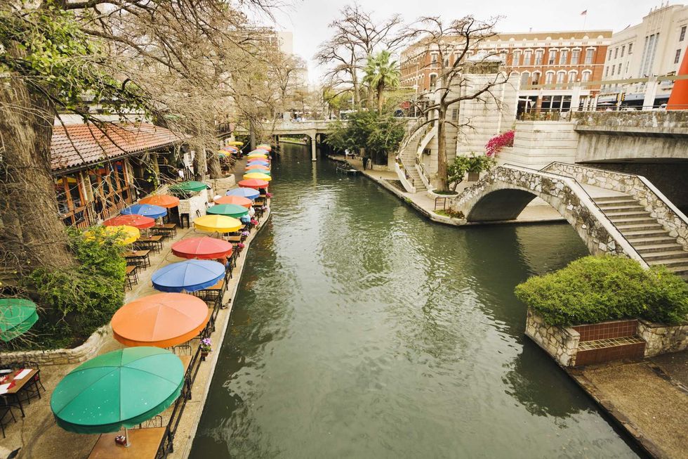 Colorful umbrellas line a riverside walkway with a stone bridge overhead.
