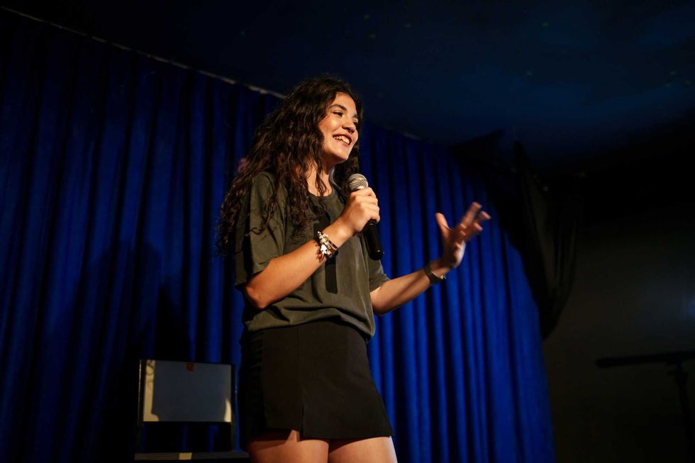 Comedian performs on stage with a microphone against a blue curtain background.