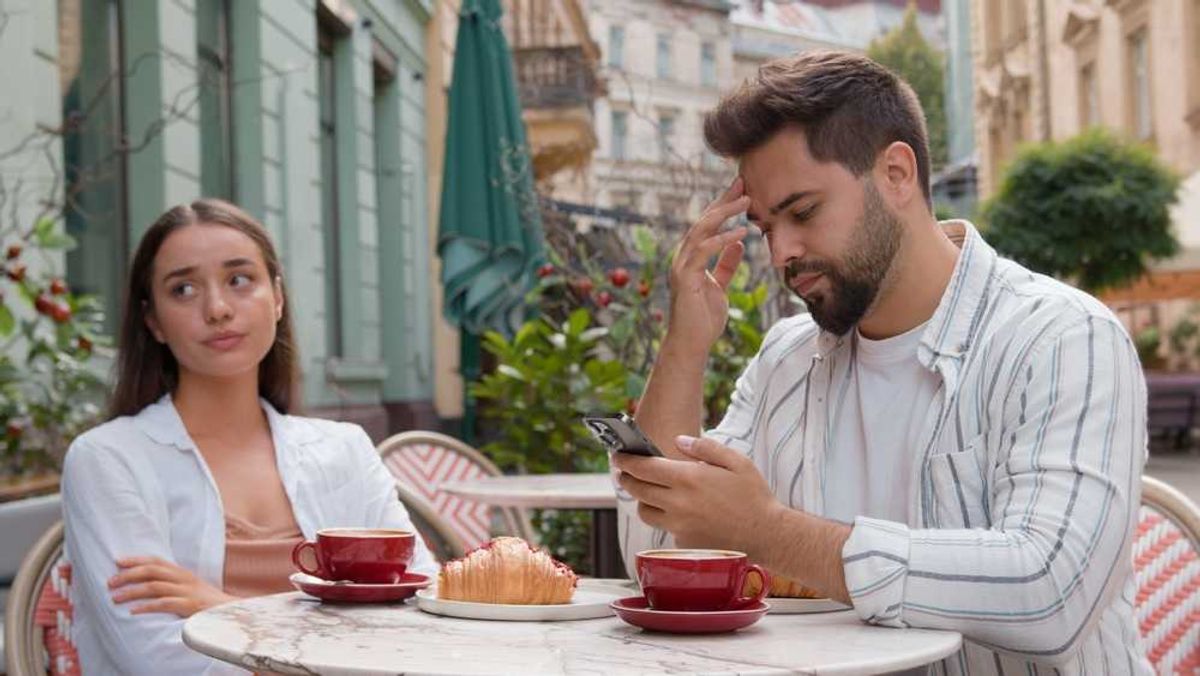 Couple at cafe, woman looks frustrated, man focused on phone, coffee and pastry on table.