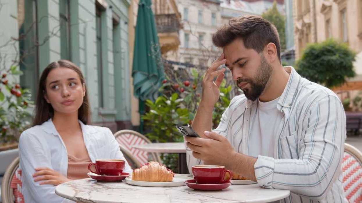 Couple at cafe, woman looks frustrated, man focused on phone, coffee and pastry on table.