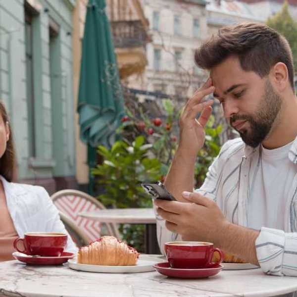 Couple at cafe, woman looks frustrated, man focused on phone, coffee and pastry on table.