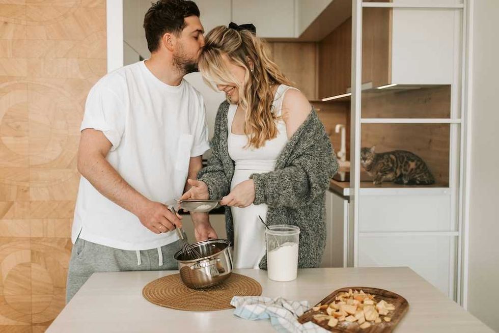Couple cooking together in kitchen; man kisses woman, cat sits on counter nearby.