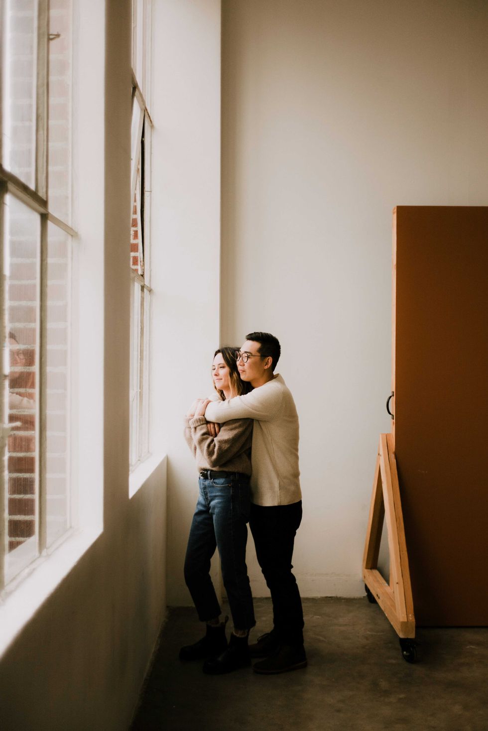 Couple embracing and looking out a large window in a softly lit room.