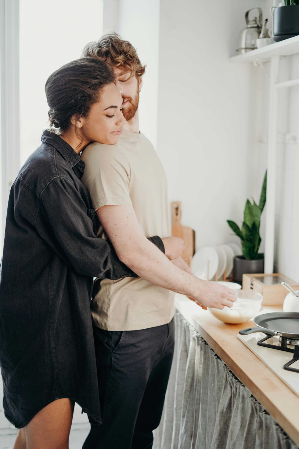 Couple embracing in kitchen, with one cooking and the other hugging from behind.