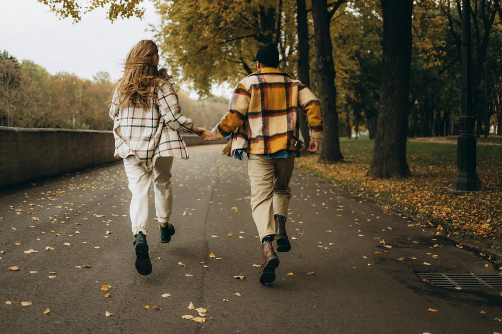 Couple holding hands, running on a tree-lined path with scattered autumn leaves.