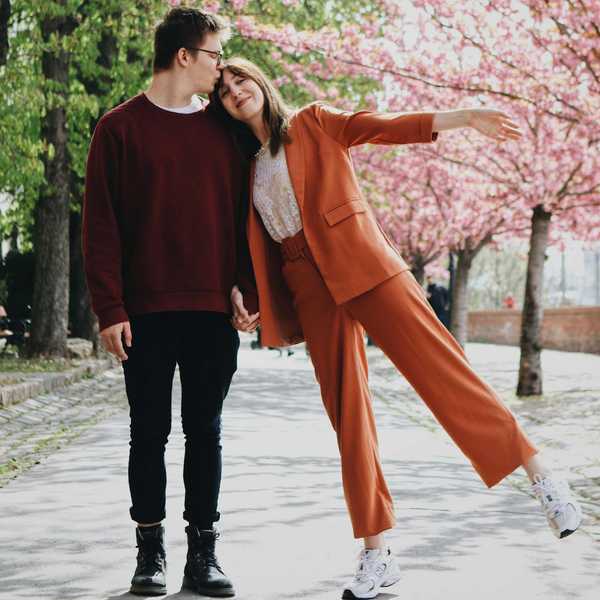Couple holding hands under cherry blossoms; woman playfully leans and lifts a leg.