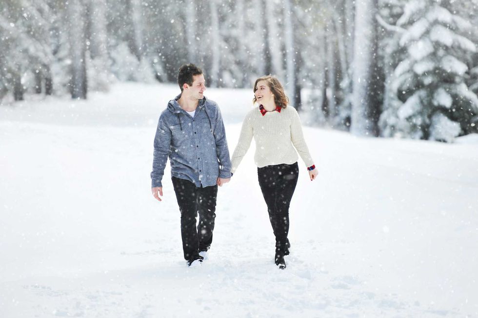 Couple holding hands, walking in snow-covered forest during snowfall.
