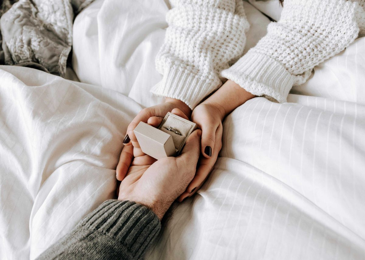 Couple holding hands with a ring box on a white bedspread.