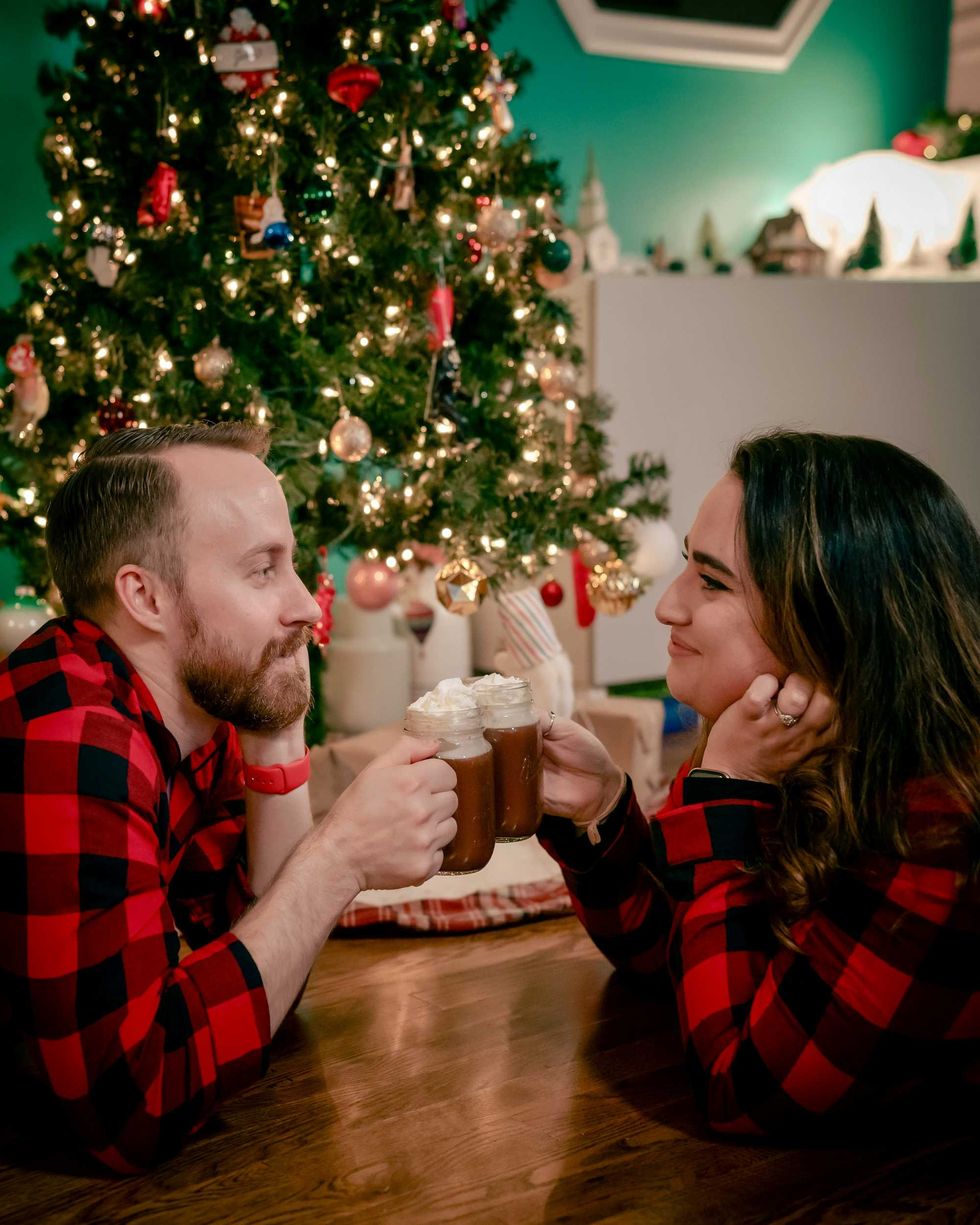 Couple in matching pajamas enjoying hot cocoa by a decorated Christmas tree.
