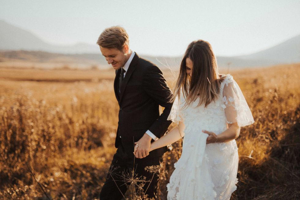 Couple in wedding attire walking hand in hand through a golden field at sunset.