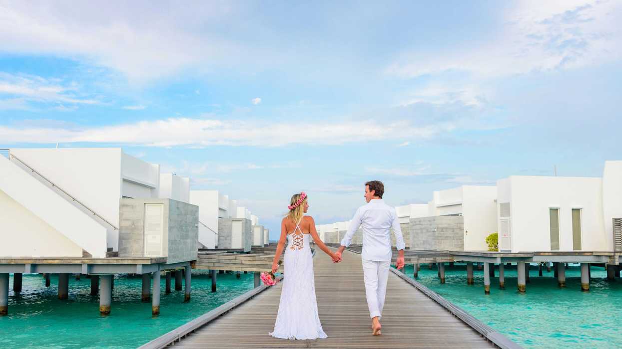 Couple in white attire holding hands on a wooden pier, surrounded by overwater villas.