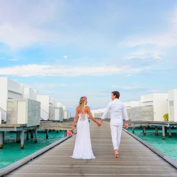 Couple in white attire holding hands on a wooden pier, surrounded by overwater villas.
