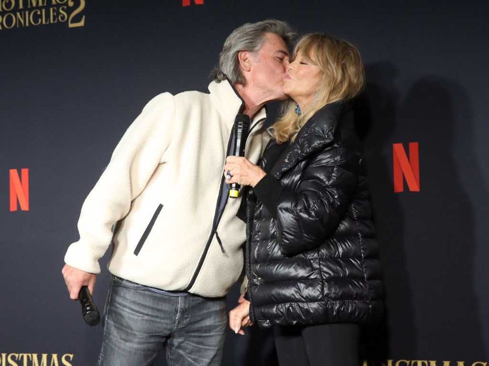 Couple kissing at Netflix event, holding microphones, wearing jackets.