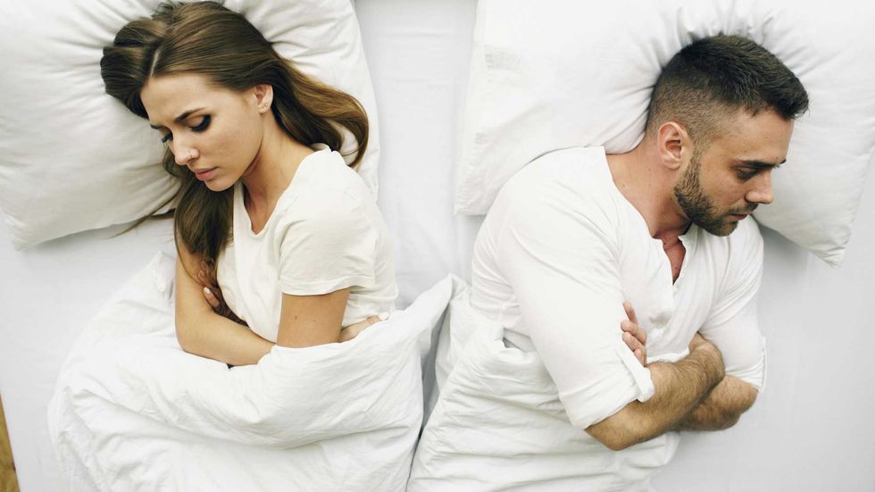 Couple lying back-to-back in bed, both looking upset with arms crossed.