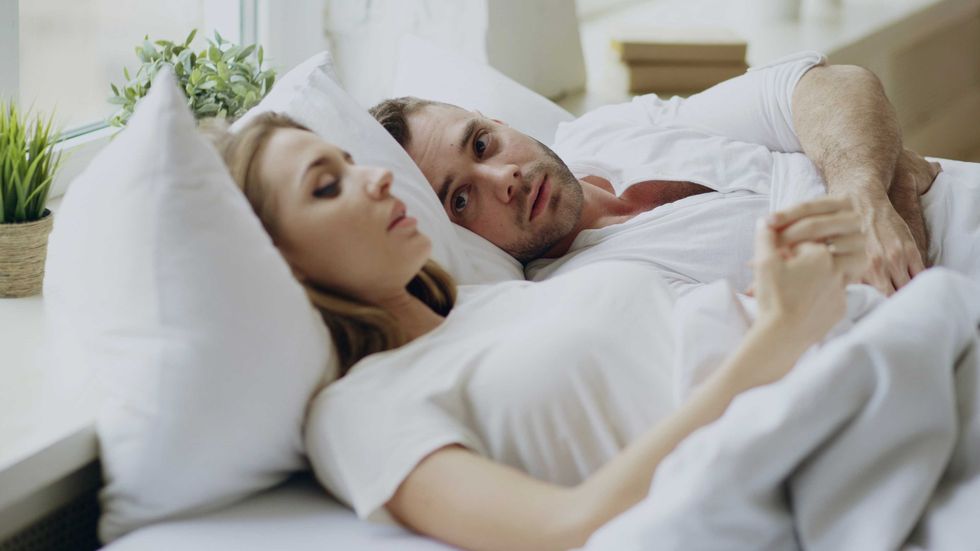 Couple lying in bed, man awake and woman sleeping, with plants in the background.