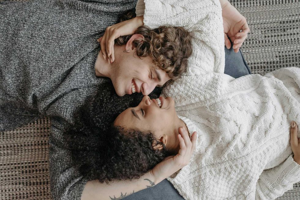 Couple lying on a rug, smiling at each other, wearing cozy sweaters.