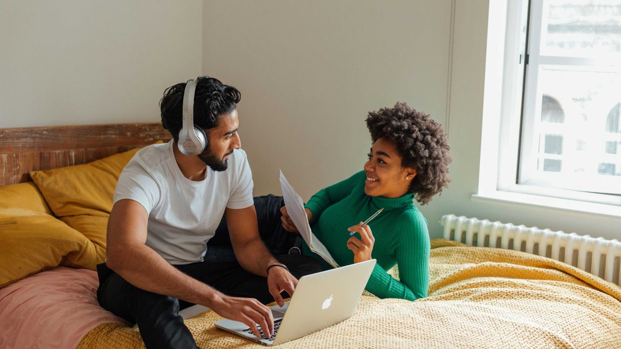 Couple on bed, man with laptop and headphones, woman holding papers and smiling at him.