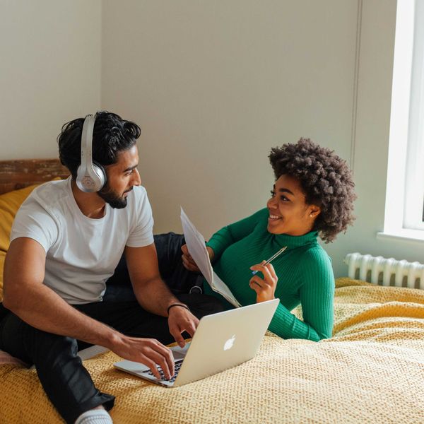 Couple on bed, man with laptop and headphones, woman holding papers and smiling at him.
