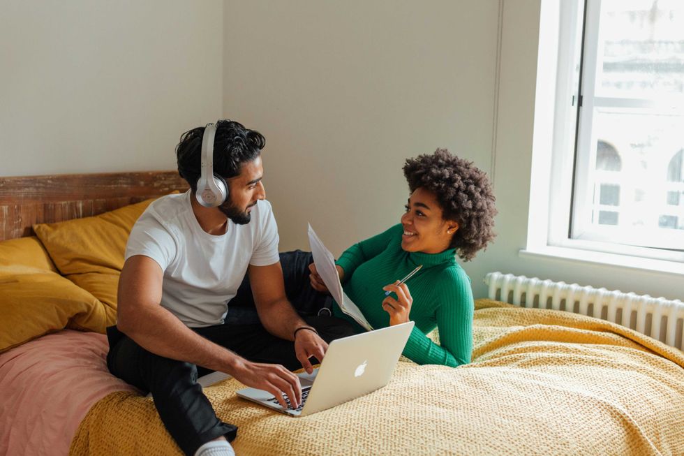Couple on bed, man with laptop and headphones, woman holding papers and smiling at him.