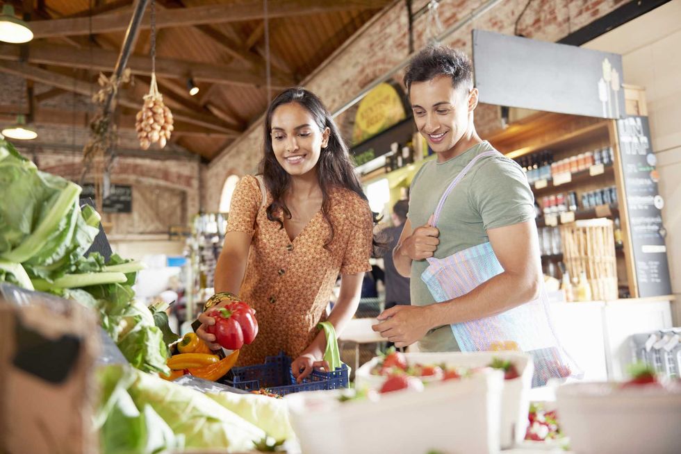 Couple shopping for produce at a farmers market, holding a red pepper and reusable bag.