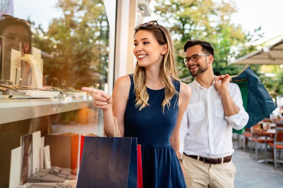 Couple shopping, woman pointing at jewelry display, man holding shopping bags and umbrella.