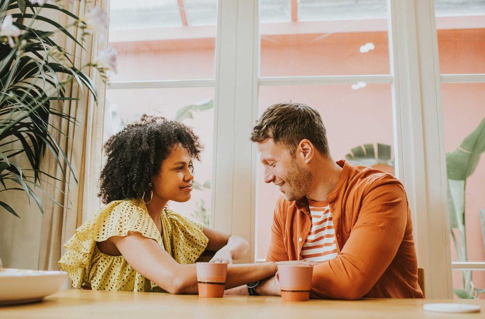 Couple sitting at a table, smiling at each other with two mugs in front of them.