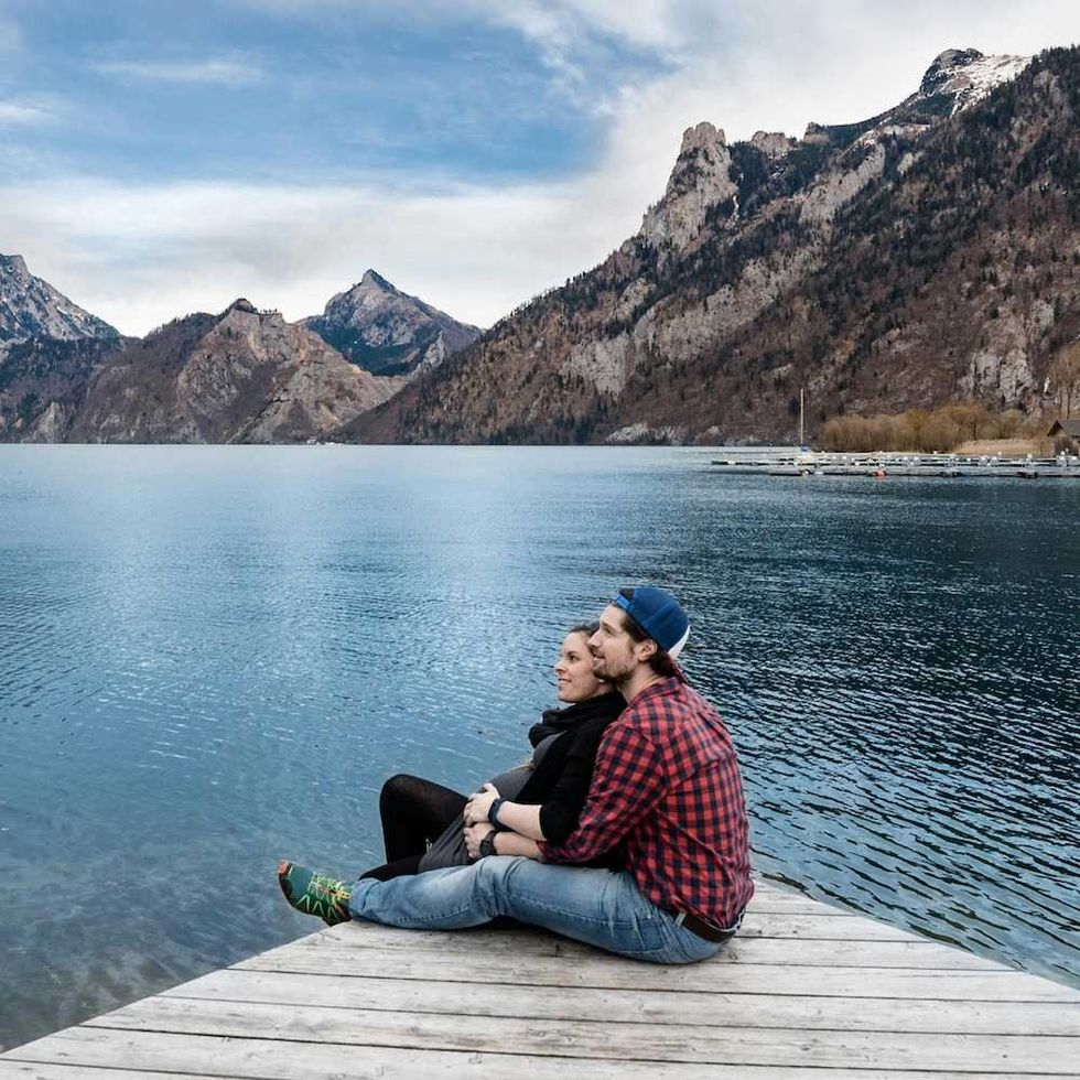 Couple sitting on a dock by a lake, surrounded by mountains under a blue sky.