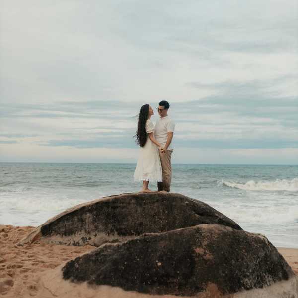 Couple standing on a rock by the ocean, holding hands, with a cloudy sky above.