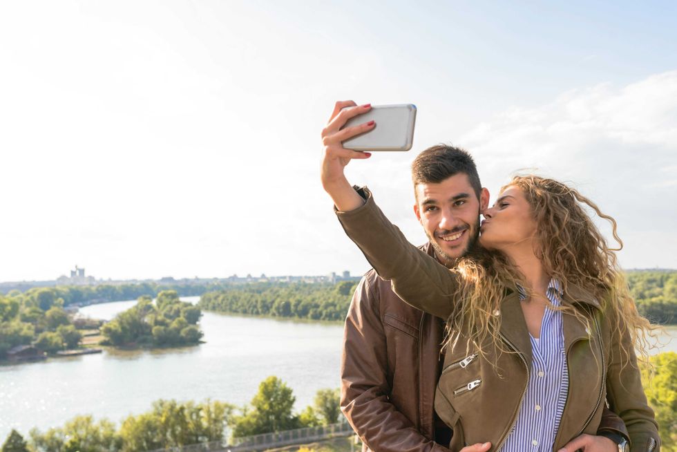 Couple taking a selfie by a river; woman kisses man's cheek.