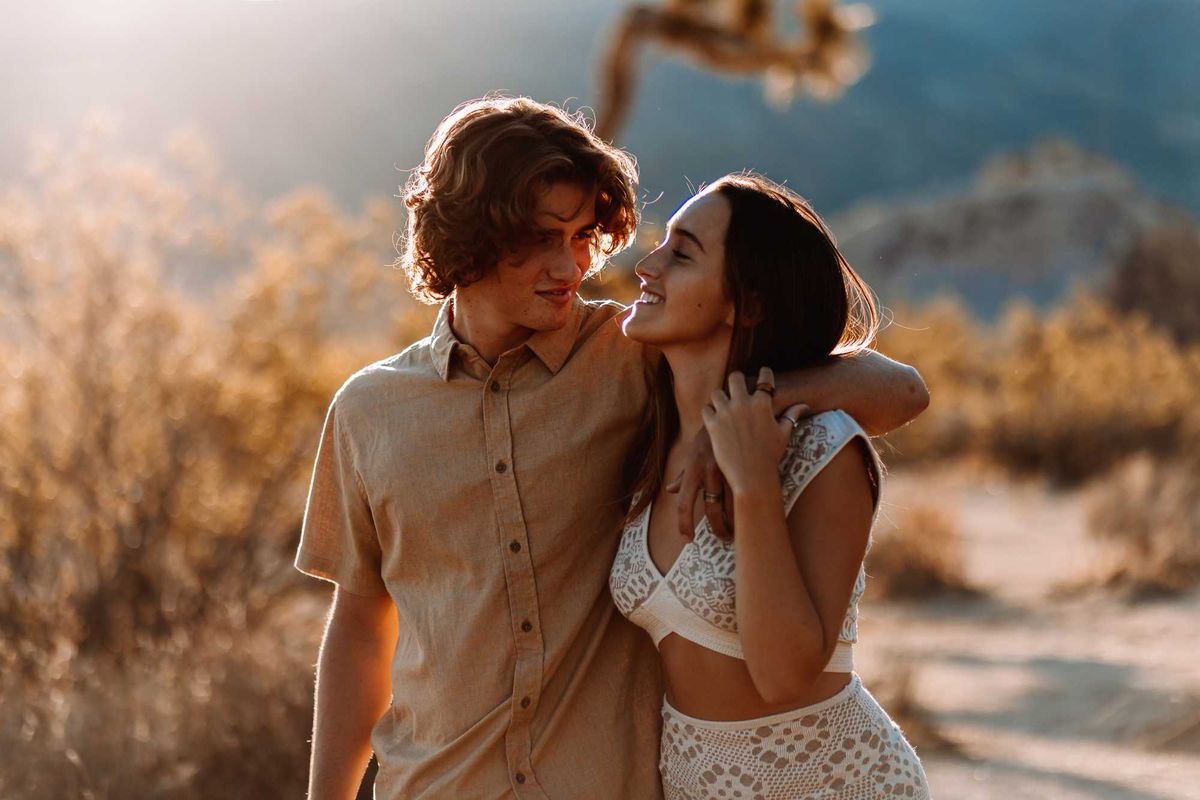 Couple walking affectionately on a sunlit path in a natural, desert-like setting.