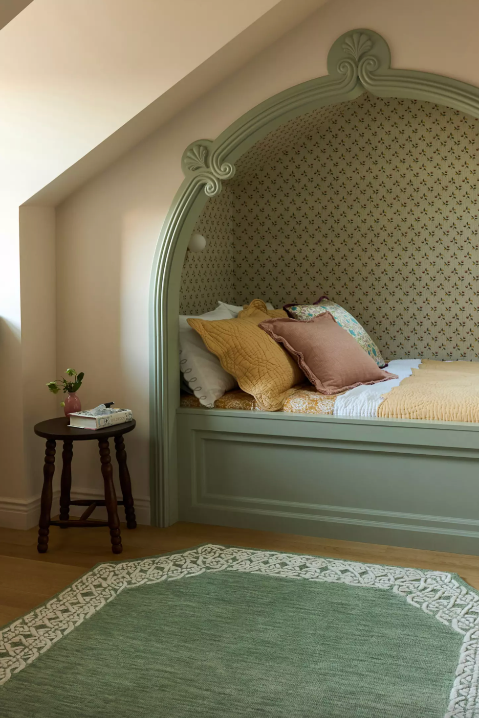 Cozy alcove bed with pillows, patterned wallpaper, round side table, and green rug.