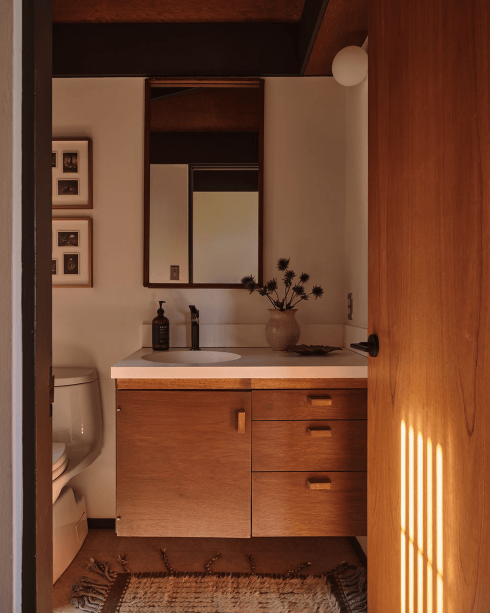 Cozy bathroom with wood vanity, mirror, and sunlight hitting the door.