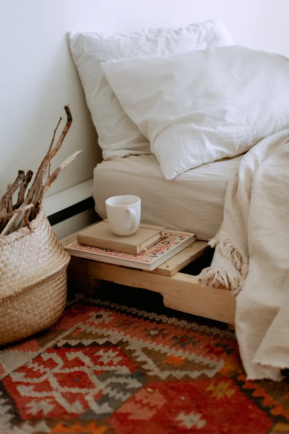 Cozy bedroom nook with white bedding, a mug on books, and a woven basket on a patterned rug.