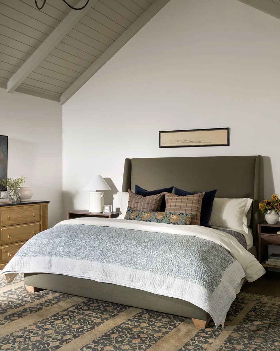 Cozy bedroom with a taupe bed, patterned bedding, wooden dresser, and floral decor.