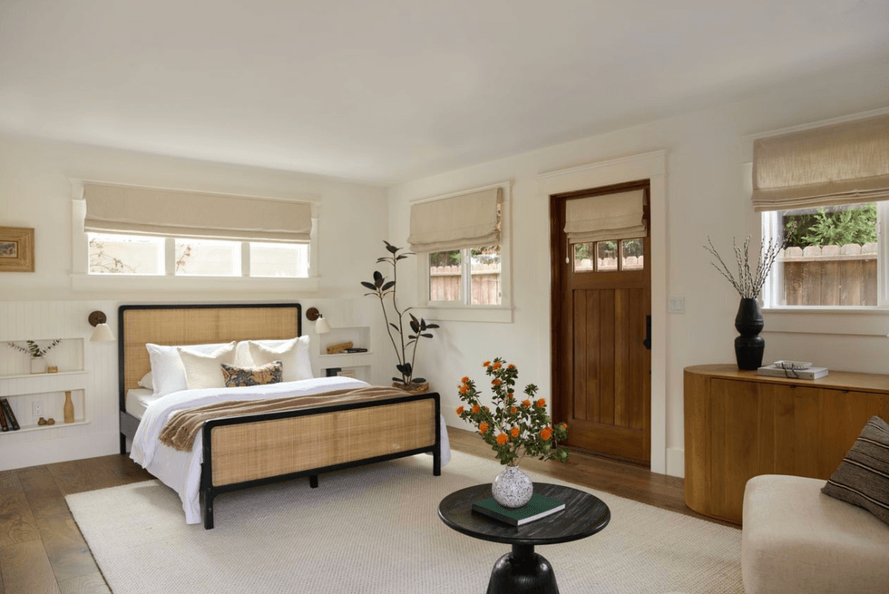 Cozy bedroom with a wooden bed, plants, and natural light through shaded windows.