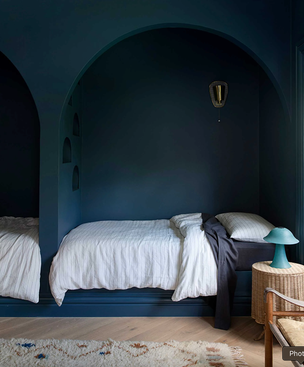 Cozy bedroom with arched alcove beds, dark walls, and wicker nightstand with a teal lamp.