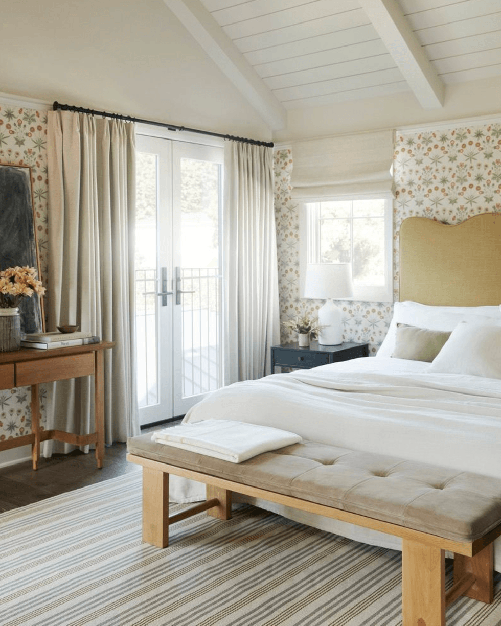 Cozy bedroom with floral wallpaper, beige accents, and a tufted bench by a sunlit window.