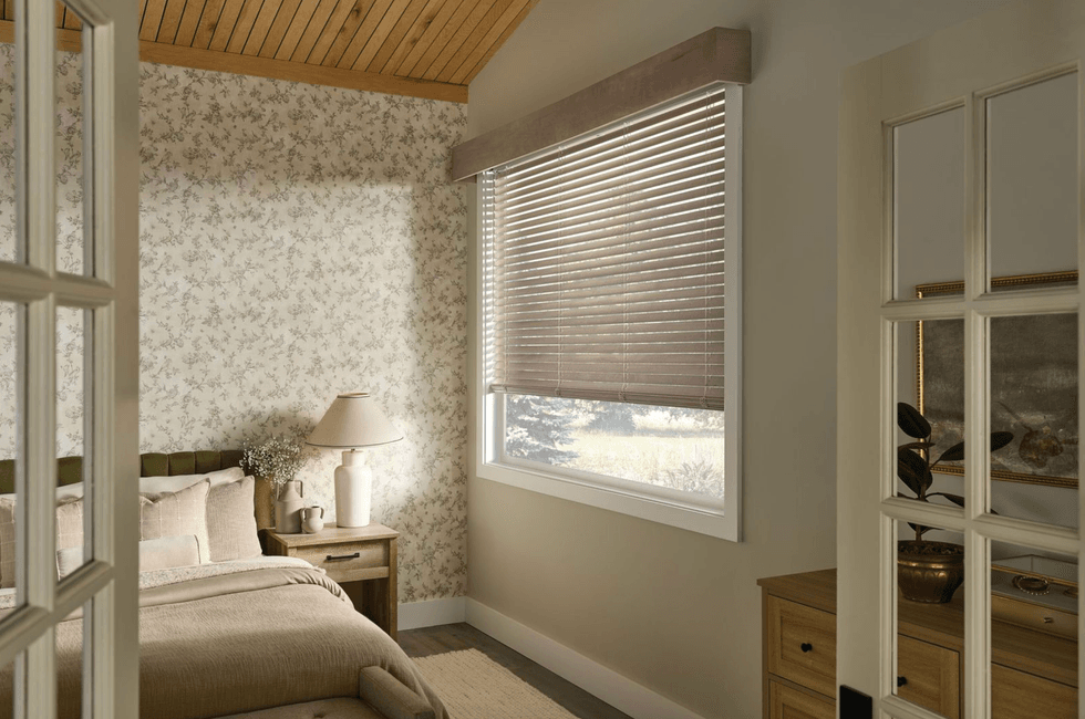 Cozy bedroom with floral wallpaper, wooden ceiling, and sunlight through window blinds.