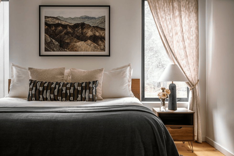 Cozy bedroom with landscape art, neutral bedding, and window light filtered by sheer curtains.
