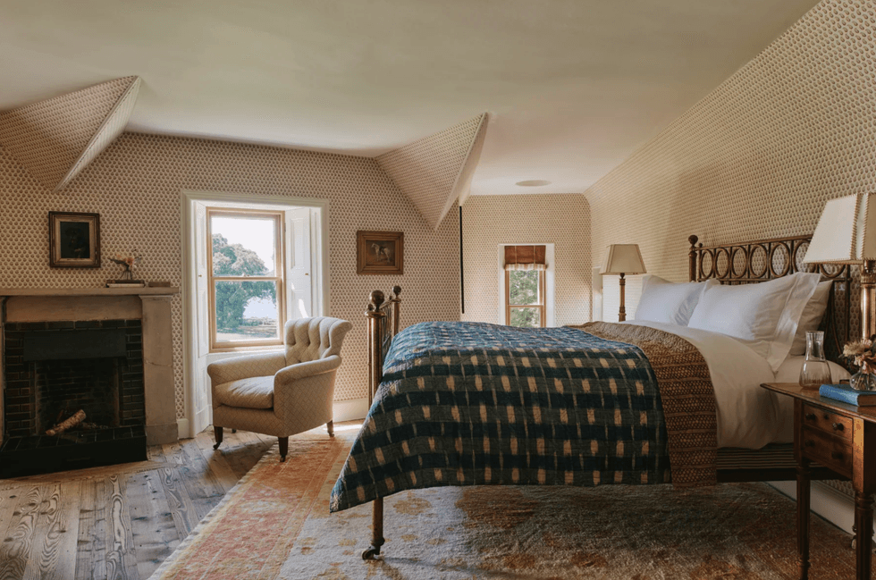 Cozy bedroom with patterned wallpaper, bed, armchair, fireplace, and a window with a scenic view.