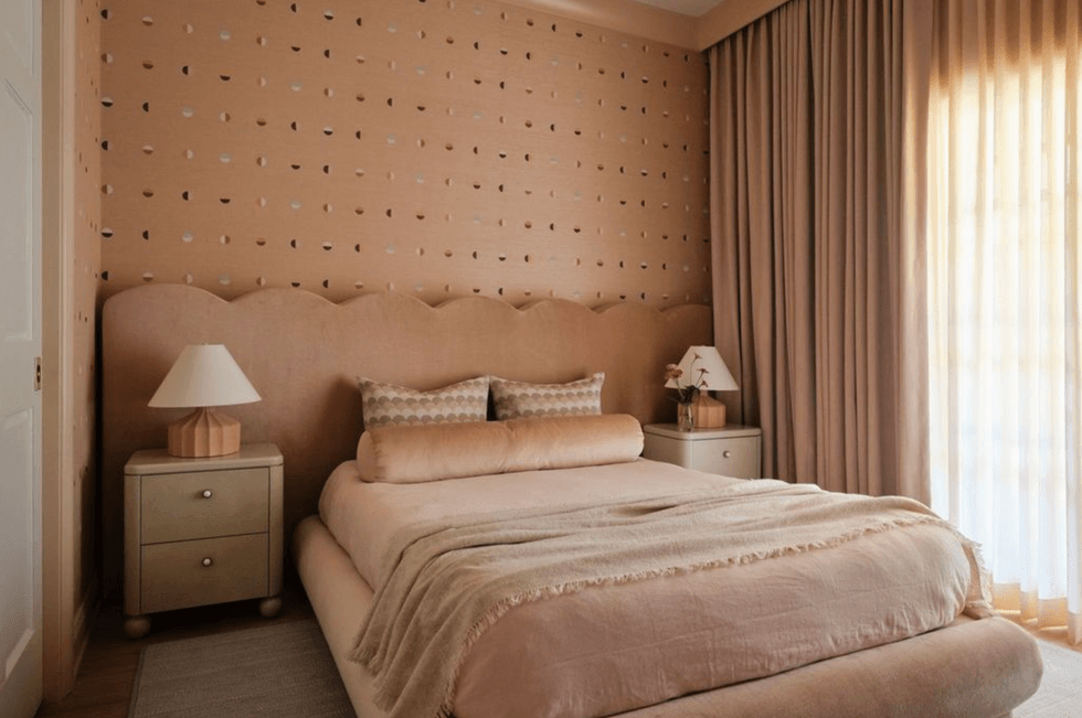 Cozy beige bedroom with a patterned wall, plush bed, and soft lighting from large curtains.