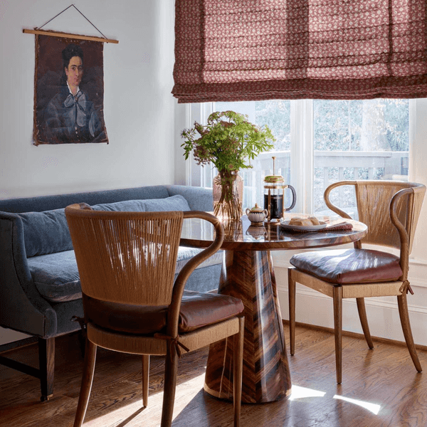 Cozy dining nook with blue sofa, wooden chairs, pendant light, artwork, and vase on table.