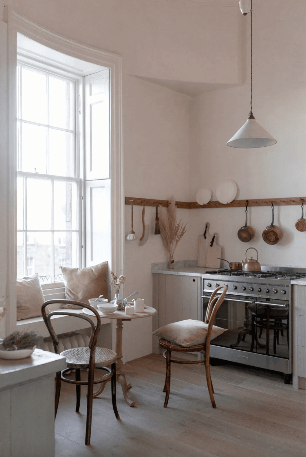 Cozy kitchen with vintage decor, wooden chairs, and a small table by a large window.