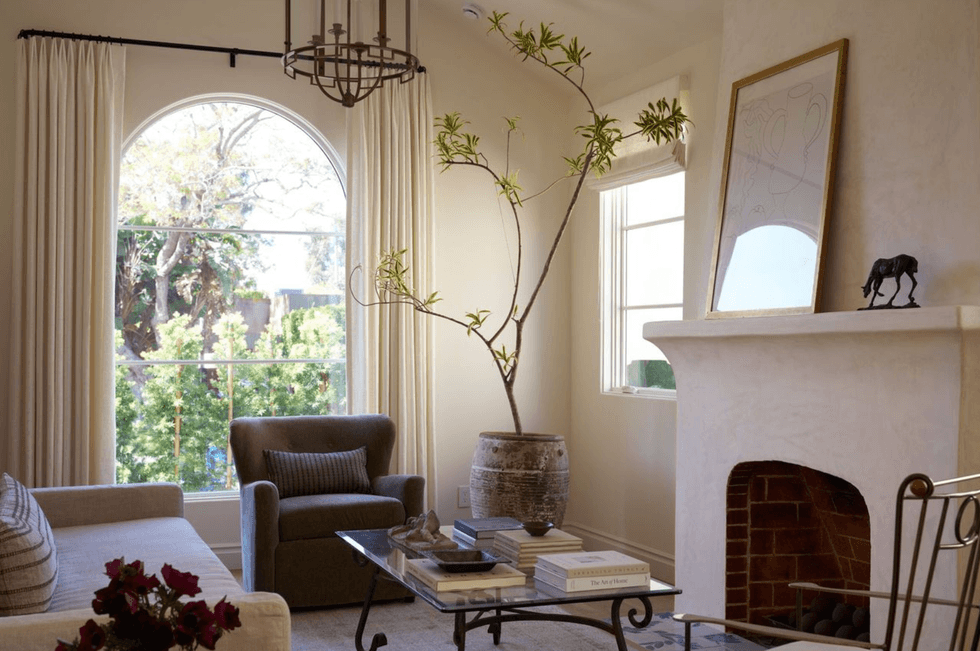 Cozy living room with large arched window, armchairs, and fireplace adorned with art.