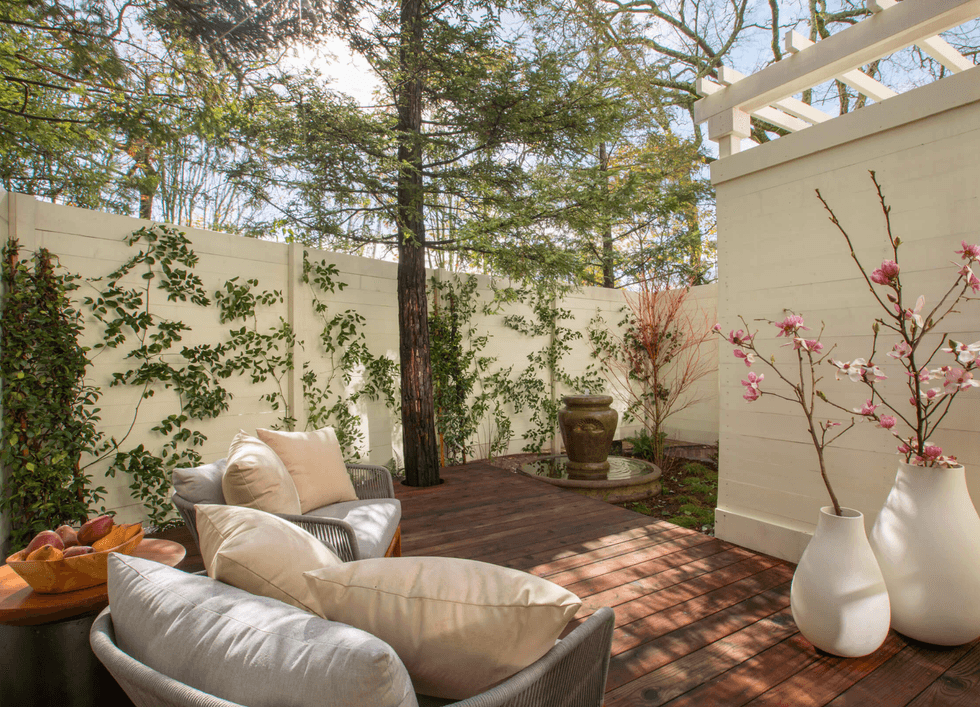 Cozy patio with chairs, potted plants, flowers, and a small fountain against a white fence.