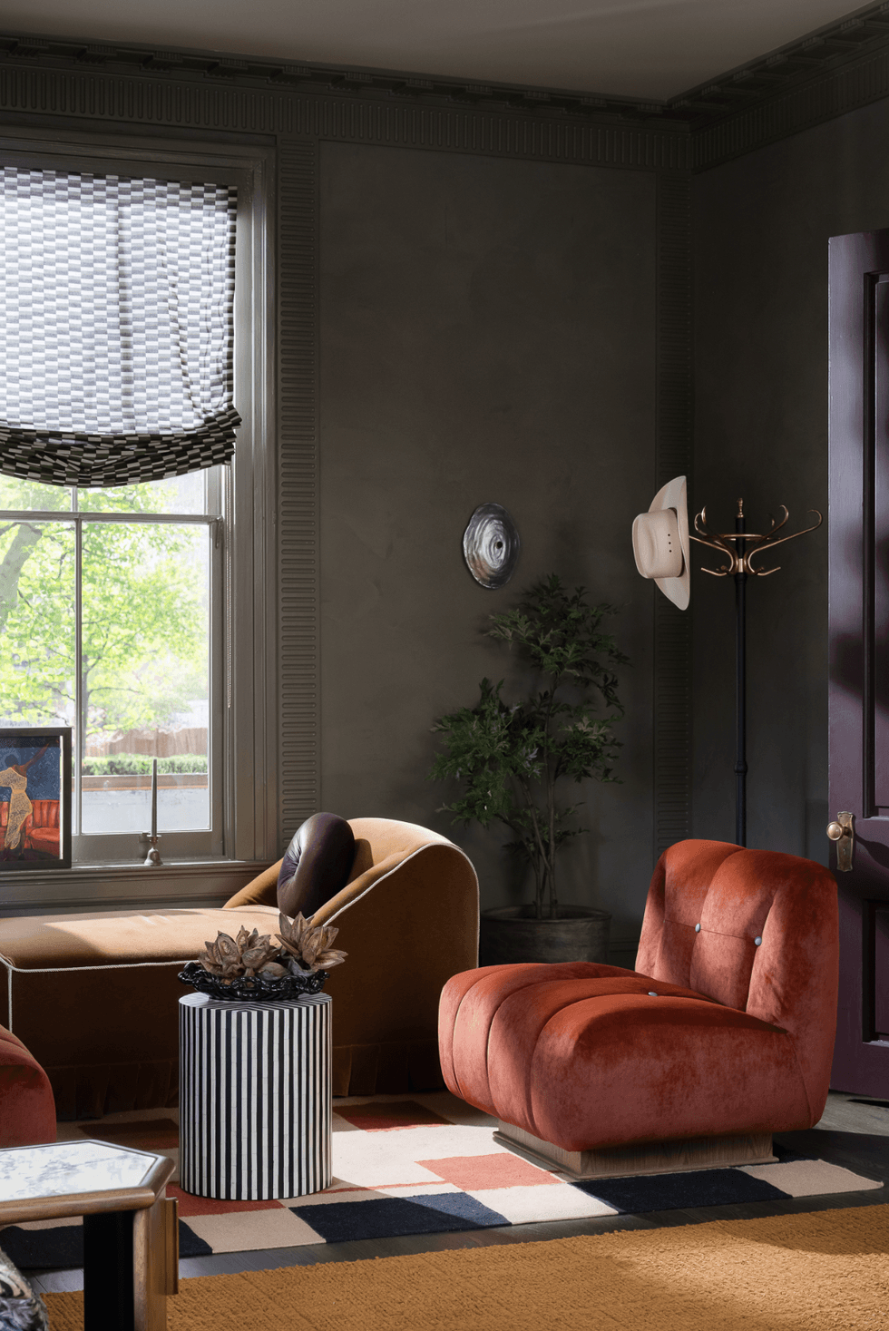 Cozy room with velvet chairs, striped table, and wall art. Large window shows greenery outside.
