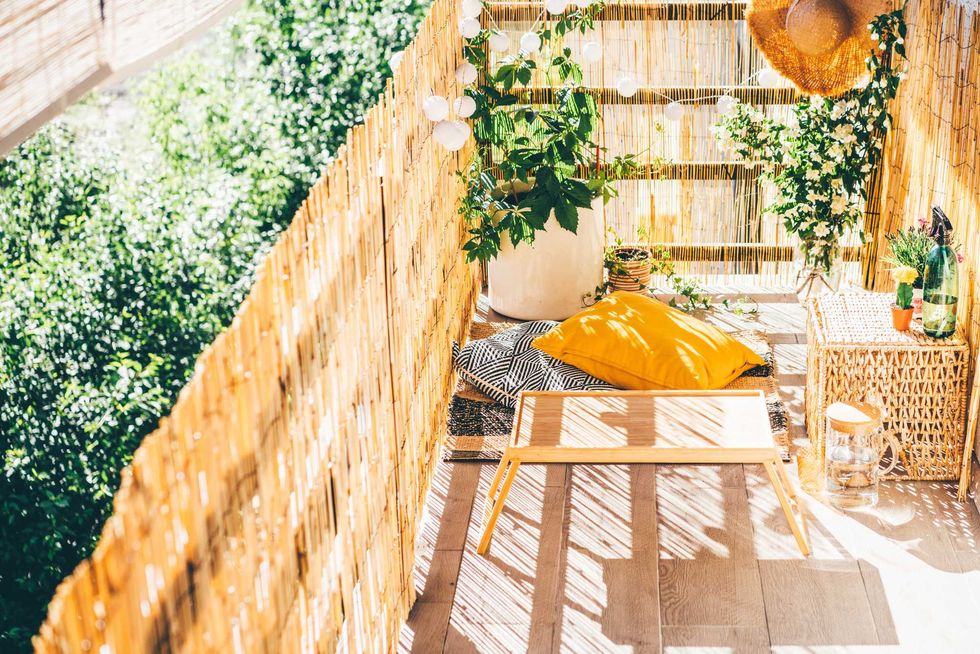 Cozy small balcony with plants, rattan furniture, cushions, and warm sunlight.