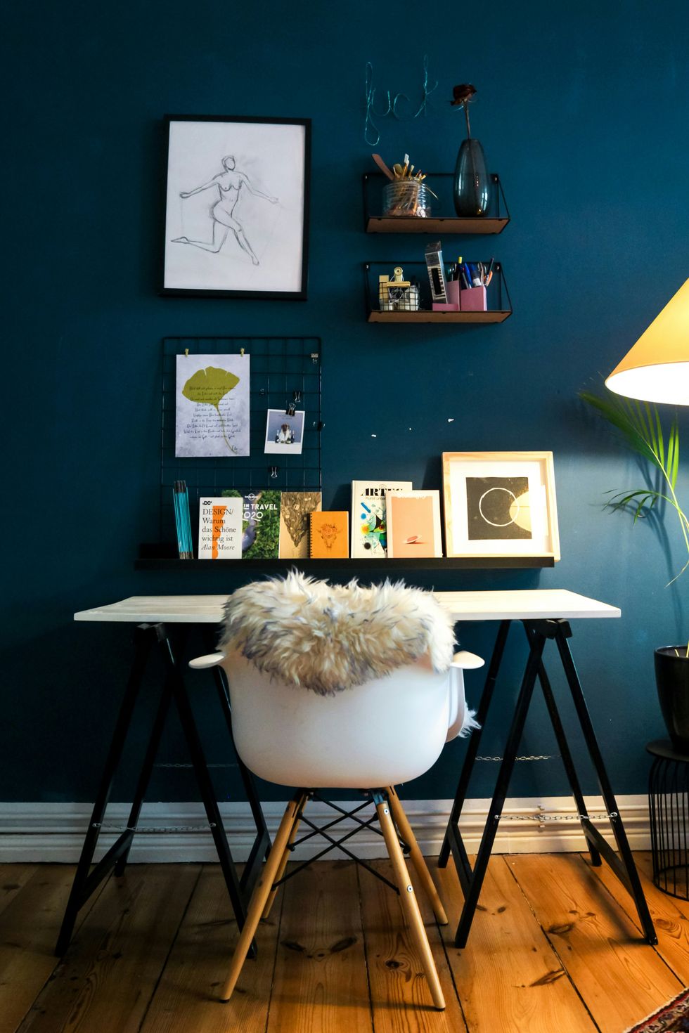 Cozy workspace with art, shelves, and a fur-topped chair against a teal wall.